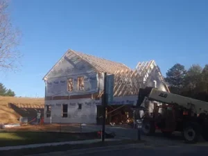 Exterior view of a house under construction, wrapped in house wrap with exposed roof trusses, next to a yellow JLG telehandler