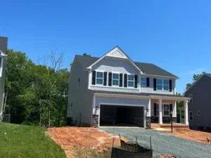 Newly constructed two-story suburban house with light gray siding, black shutters, and a stone-accented garage entrance under a bright blue sky