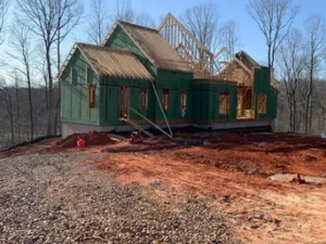 Residential house under construction on a red dirt lot, with the lower walls wrapped in green sheathing