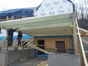Two construction workers standing on temporary scaffolding below a newly installed, heavy timber balcony