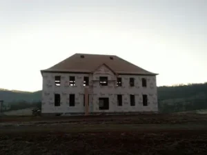 house under construction on a muddy, open lot, wrapped entirely in white house wrap with a partially covered roof
